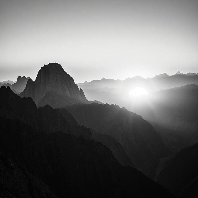 Uma cena tranquila do sol se pondo sobre uma cadeia de montanhas, lançando um suave brilho sobre o vale enevoado abaixo. A paleta em preto e branco realça a atmosfera serena desta paisagem.