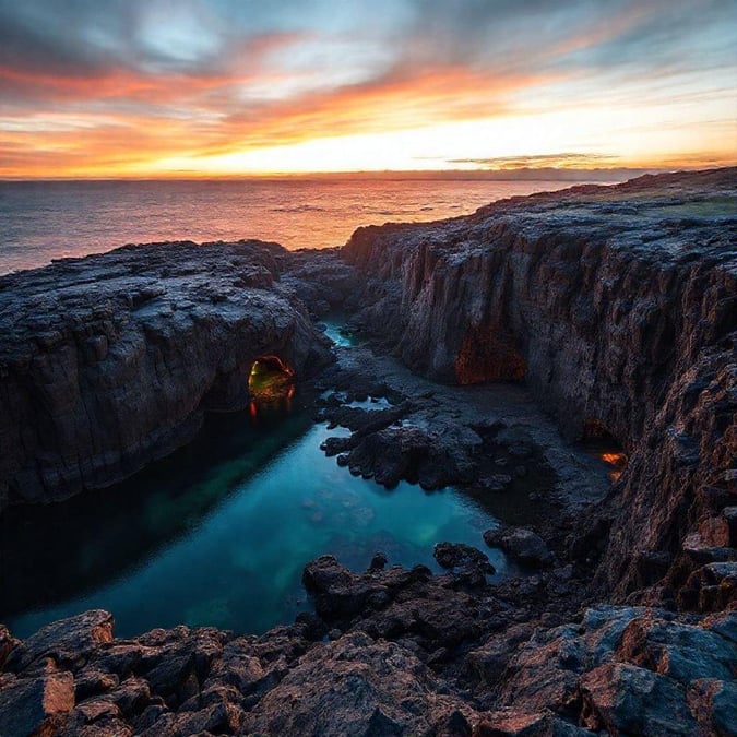 A serene view of the sun dipping below the horizon at a rocky cove, with warm hues illuminating the tranquil waters.