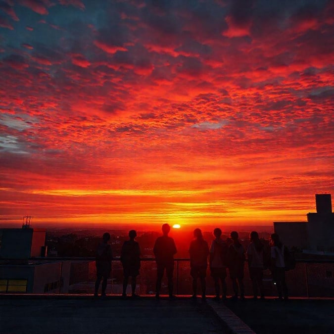 Ryhmä ystäviä kokoontui katolle katsomaan, kuinka taivas täyttyy eloisilla punaisen ja oranssin sävyillä auringonlaskun aikana. Kaupungin rakennusten siluetti näkyy upean taustan edessä. Koulun paluun henki vangittuna.